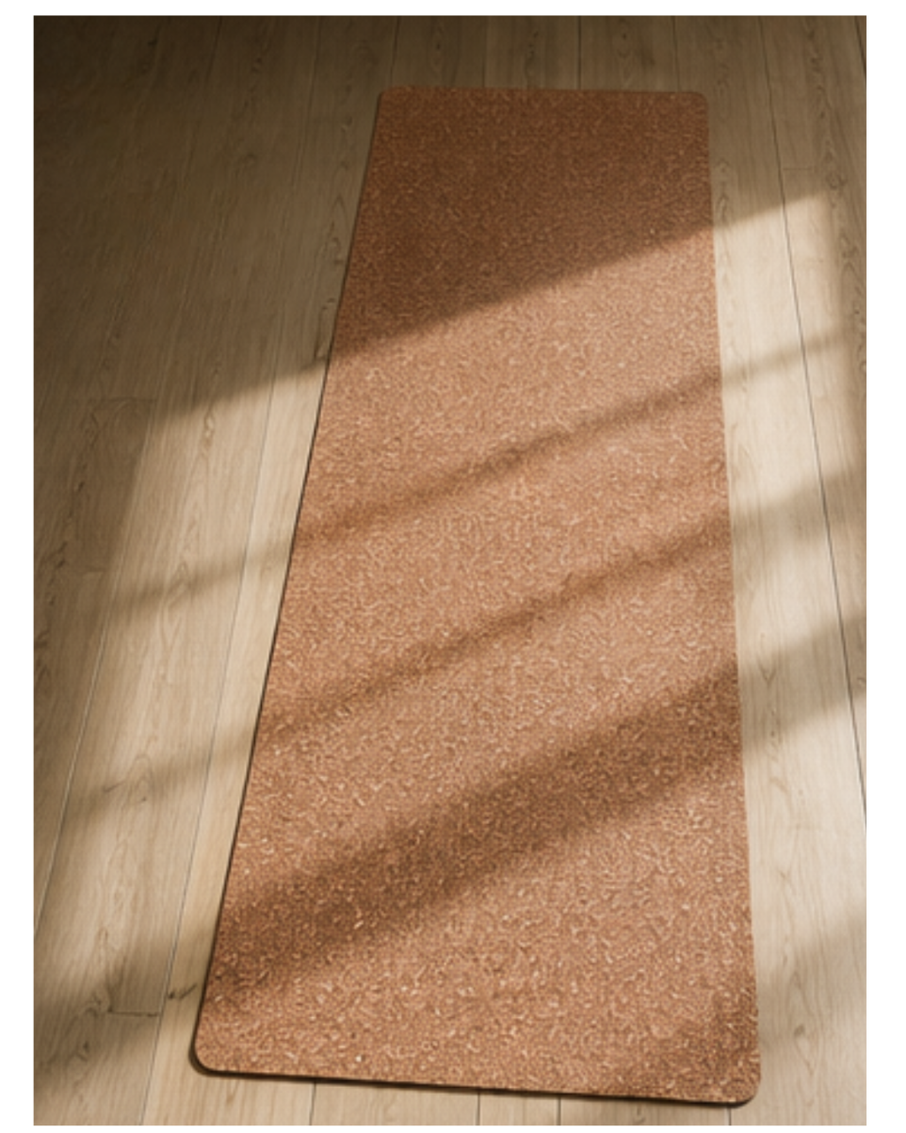 A rectangular cork yoga mat with rounded corners lies on a light wood floor, illuminated by soft diagonal sunlight casting shadows.