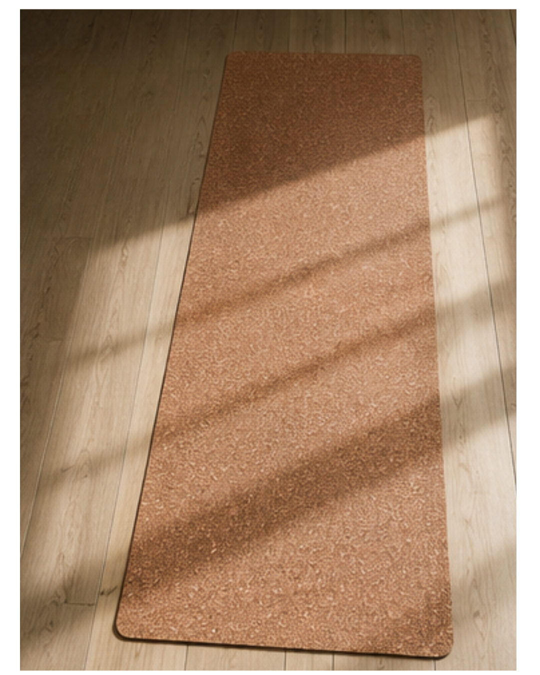 A rectangular cork yoga mat with rounded corners lies on a light wood floor, illuminated by soft diagonal sunlight casting shadows.