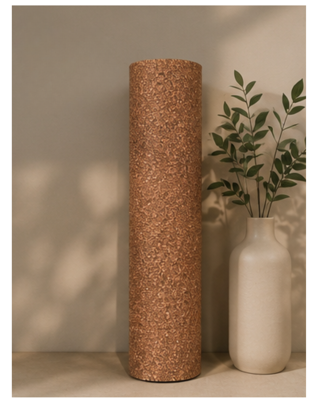 A rolled cork yoga mat, stands next to a white ceramic vase with green leaves, set against a warm beige wall with soft leaf shadows.